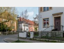 Straßenszene in einem Wohngebiet mit gepflastertem Gehweg und kleinen Vorgärten. Links eine Straßenecke mit einem grünen Laternenpfahl und Straßenschildern, im Hintergrund farbige Reihenhäuser mit roten Dächern, rechts ein weißes Haus mit Veranda und Tor.