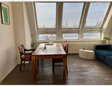 Bright attic-style living and dining area with slanted ceiling windows fitted with blinds, overlooking rooftops. A wooden dining table with chairs sits in the center, with a blue sofa and curved white side table beside a radiator and potted plants.