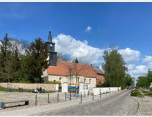 Historische Dorfkirche mit hellem Putz, rotem Ziegeldach und einem dunklen Kirchturm mit Uhr, umgeben von Bäumen und einer niedrigen Steinmauer; im Vordergrund eine gepflasterte Straße mit Parkbänken und Fahrrädern.