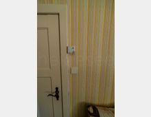 Innenraum mit beige gestrichener Holztür und dunkler Klinke neben einer Wand mit gelb-grau gestreifter Tapete. An der Wand sind ein Lichtschalter und ein kleines Bediengerät montiert; unten ist die Ecke eines gepolsterten Möbels sichtbar.