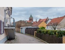 Schmale gepflasterte Gasse mit Wohnhäusern, Holzzäunen und Garagen. Im Hintergrund ist ein Kirchturm mit grünem Dach und roten Backsteinen sichtbar, umgeben von weiteren Häusern mit roten Ziegeldächern.