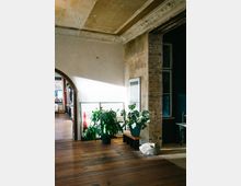 Wohnraum mit rustikalem Charakter, geprägten Holzfußböden, einer freiliegenden Backsteinwand und verzierten Stuckelementen an der Decke. Im Vordergrund stehen Topfpflanzen auf einer Bank, und ein Bogen führt in einen weiteren Raum, der teilweise sichtbar ist.