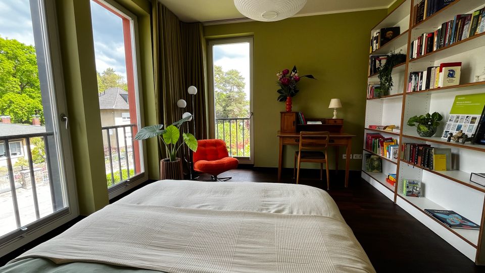 Helles Schlafzimmer mit olivgrünen Wänden, großem Bett im Vordergrund und bodentiefen Fenstern mit Balkonblick ins Grüne. Rechts stehen ein langes Bücherregal und ein kleiner Schreibtisch; im Hintergrund ein roter Sessel, Pflanzen und eine runde Deckenlampe.