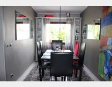 Esszimmer mit einer zentralen, schwarz gedeckten Esstischgarnitur und vier schwarzen Stühlen sowie einem roten Stuhl. Der Raum ist modern gestaltet, mit grauen Wänden, großen Spiegeln an den Seiten, einem Teppich unter dem Tisch und einem Fenster mit Ausblick ins Grüne.