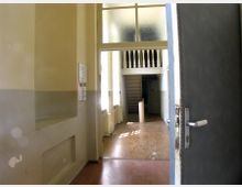 Eine geöffnete Tür führt in einen Flur mit cremefarbenen Wänden und einem Holzboden; im Hintergrund ist eine weiße Treppe und ein Zwischengeschoss mit einer Balustrade sichtbar. Das Licht fällt durch große Fenster, die teils mit milchigem Glas versehen sind.