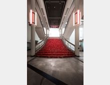 Treppe mit rotem Teppich in einem modernen Kinogebäude, flankiert von weißen Säulen und Neonleuchten; ein Schild über der Treppe weist auf die Kinos 6 bis 10 hin. Die Decke ist dunkel gestaltet, und helle Wände sowie große Fenster lassen Tageslicht in den Raum.