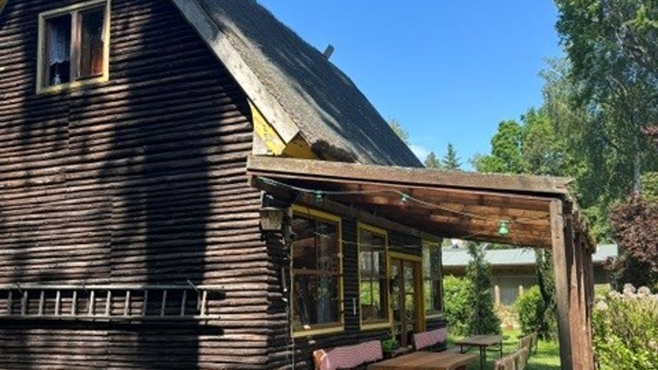Holzhaus mit steilem Satteldach und kleinem Dachfenster, daneben eine überdachte Veranda mit Sitzbank und Tisch. Das Gebäude steht in einem grünen Garten mit Rasen und Bäumen unter klarem blauem Himmel.