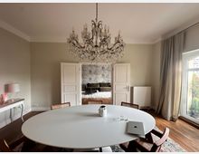 Helles Esszimmer mit einem runden, weißen Tisch und Stühlen aus Holzgeflecht. Ein großer Kronleuchter hängt von der Decke, und durch eine offene Doppeltür ist ein angrenzender Raum mit Bett und gemusterter Wandtapete sichtbar; rechts befindet sich ein Fenster mit Gardinen.