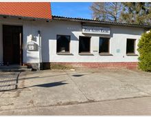 Ein einstöckiges Gebäude mit weißer Fassade und gemauertem Sockel, auf dem ein Schild mit der Aufschrift „Zur Alten Eiche“ angebracht ist. Links befindet sich eine Eingangstür unter einem roten Ziegeldach, während der Vorplatz aus Beton besteht und rechts ein großer Nadelbaum sichtbar ist.