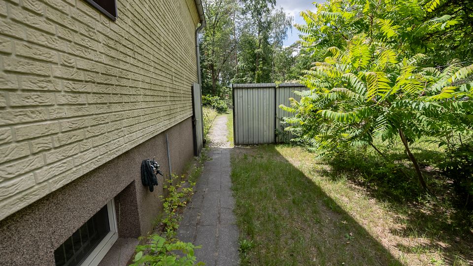 Schmaler gepflasterter Weg entlang der Seitenwand eines Wohnhauses mit hell gestrichener Ziegel-Fassade und Kellerfenstern. Rechts grenzt ein grasbewachsener Gartenstreifen mit dichtem Strauch- und Baumbestand an; am Ende steht ein graues Metalltor.
