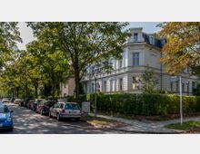 Straßenszene in einem Wohngebiet mit einer historischen Stadtvilla aus der Gründerzeit auf der rechten Seite, umgeben von gepflegten Hecken und Bäumen. Am Straßenrand sind parkende Autos zu sehen, während die Straße von hohen, belaubten Bäumen gesäumt ist.