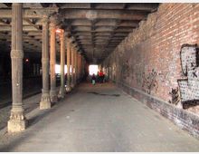 Ein schmaler, überdachter Durchgang mit Ziegelwänden, verziert mit Graffiti, links flankiert von einer Reihe dekorativer Metallsäulen. Im Hintergrund ist ein heller Ausgang sichtbar, während Personen den Tunnel durchqueren.