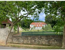 Ein zweistöckiges weißes Gebäude mit rotem Ziegeldach und mehreren Fenstern, umgeben von Bäumen und einer gepflegten Gartenanlage. Im Vordergrund eine hohe Steinmauer mit grünem Zaun, flankiert von einem Torbogen aus Stein mit Ziegeldach.
