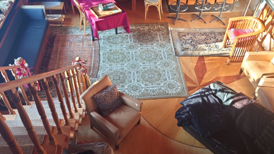 Wohnraum mit offener Gestaltung, Holzboden und verschiedenen Ebenen. Die Einrichtung umfasst einen Esstisch mit Stühlen, eine Sitzecke mit Sesseln und Sofas, Teppiche und eine hölzerne Treppe, die zu einem erhöhten Bereich führt.