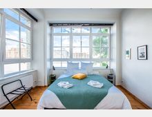 Helles Schlafzimmer mit großem Bett in der Mitte, dekoriert mit weißer und grüner Bettwäsche sowie einem gelben Kissen, flankiert von Nachttischen mit Lampen. Im Hintergrund große Fenster mit Blick auf eine urbane Landschaft und Bäume, Holzboden und weiße Wände mit minimalistischer Dekoration.