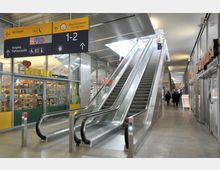 Einkaufszentrum mit zwei Rolltreppen, die nach oben führen, flankiert von Glasfassaden, Geschäften und einem gelben Wegweiser mit Informationen zu Ausgängen und Bereichen wie Raucherzone und WC.