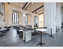 Innenansicht eines modernen Kirchengebäudes mit hohen, hellen Wänden und großen, bogenförmigen Fenstern. Im oberen Bereich befinden sich Sitzgruppen mit runden Tischen, während im Hintergrund ein beleuchtetes Kreuz und weitere Sitzreihen sichtbar sind.