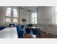 Ein heller Büro- oder Besprechungsraum mit hohen Fenstern und weiß gestrichenen Wänden. Der Raum ist mit einem großen Tisch, blauen Stühlen und einem Schreibtisch mit Computer ausgestattet, außerdem sind Wanddekorationen und ein Pflanzenständer zu sehen.