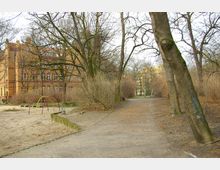 Ein kleiner Park mit einem Sandspielplatz, auf dem ein Klettergerüst aus roten und gelben Metallbögen steht. Im Hintergrund befinden sich ein großes Backsteingebäude mit zahlreichen Fenstern und vereinzelt sichtbare Wohnhäuser, umgeben von kahlen Bäumen und Sträuchern.