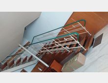 Holz-Innentreppe mit einer weißen Metallgeländerstruktur und grünem Handlauf in einem Raum mit hellblauen Wänden und Holzelementen. Im Vordergrund sind Kartons und ein Tisch sichtbar.