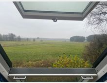 Blick aus einem geöffneten Dachfenster auf eine weite, grüne Landschaft mit Wiesen, vereinzelten Bäumen und einem Waldrand am Horizont. Der Himmel ist bewölkt, und im Vordergrund sind Zweige eines Strauches sichtbar.