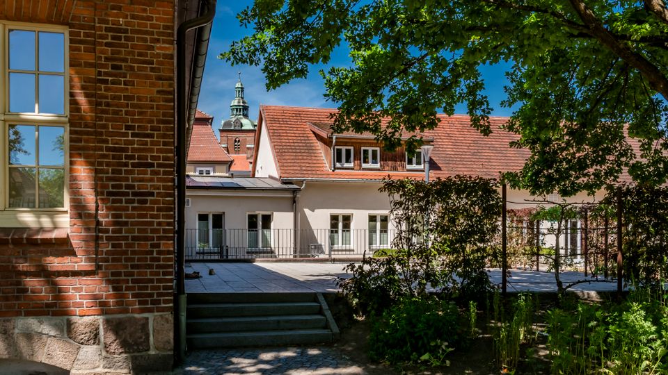 Innenhof mit gepflasterter Terrasse und kleiner Treppe zwischen einem Backsteingebäude mit großen Sprossenfenstern und einem hellen Gebäude mit roten Ziegeldächern. Rechts steht ein großer Baum mit Grünfläche und Hecken; im Hintergrund ragt ein Kirchturm über die Dächer.