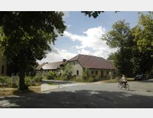 Straßenszene in einer ländlichen Umgebung mit einem Wohnhaus mit roten Dachziegeln, umgeben von Bäumen und Pflanzen. Im Vordergrund fährt eine Person auf einem Fahrrad eine asphaltierte Straße entlang.