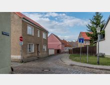 Eine kleine Kopfsteinpflasterstraße in einer Wohngegend mit einfachen zweigeschossigen Häusern, teils verputzt und teils aus Backstein, sowie roten Ziegeldächern. Links zeigt ein Straßenschild „Mauerstraße“ an, daneben stehen ein Einbahnstraßenschild und ein Laternenpfahl; im Hintergrund ist ein Gehweg und eine Grundstücksbegrenzung aus Holz.