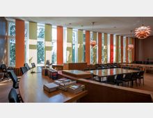 Geräumiger Sitzungssaal mit großen, bodentiefen Fenstern, verziert durch vertikale Vorhänge in Orange- und Gelbtönen. Der Raum verfügt über eine ovale Holztheke, lange Konferenztische mit schwarzen Stühlen sowie mehrere moderne, kupferfarbene Deckenleuchten.