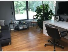 Moderner Arbeitsraum mit Holzboden, einer großen Fensterfront mit Jalousien und Blick auf grüne Bäume. Der Raum enthält einen weißen Schreibtisch mit schwarzem Bürostuhl, ein graues Sofa, ein Regal mit Büchern und Dekorationen sowie eine große Zimmerpflanze.