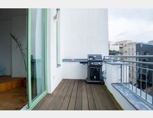 Ein Balkon mit Holzbodenbelag, einer weißen Wand und einem schwarzen Gasgrill auf der rechten Seite. Links führt eine geöffnete Tür mit grünen Rahmen ins Innere einer Wohnung, im Hintergrund sind moderne Wohngebäude zu sehen.