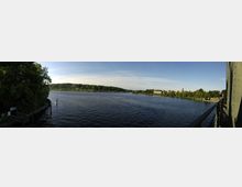 Weitläufiger Fluss mit ruhiger Wasseroberfläche, umgeben von grünen Uferbäumen und Gebäudegruppen in der Ferne; rechts im Bild ist ein Teil einer Brückenkonstruktion sichtbar.