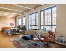 Wohnzimmer mit großen, bodentiefen Fenstern und freiliegender Ziegelwand. Der Raum ist mit Holzboden, einem Ledersessel, einem niedrigen Bücherregal, einem Teppich und gestapelten Büchern gestaltet, während viel Tageslicht einströmt.