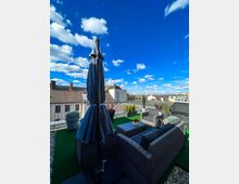 Dachterrasse mit Sitzgruppe aus grauem Rattan, grünem Kunstrasenboden, Pflanzkübeln und geschlossenem Sonnenschirm. Im Hintergrund sind Gebäude mit Ziegeldächern und ein blauer Himmel mit weißen Wolken zu sehen.