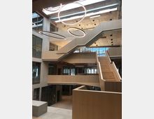 Offener, moderner Innenraum eines Gebäudes mit mehreren Ebenen und breiten Treppen aus Holz. An der Decke befinden sich runde, leuchtende Hängeelemente, während Betonwände und große Glasflächen die Architektur prägen.