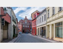 Eine kopfsteingepflasterte Straßenansicht in einer europäischen Altstadt mit bunten Gebäuden auf beiden Seiten, darunter ein rotes Ziegelgebäude mit der Aufschrift „Postamt“ im Hintergrund. Die Architektur umfasst dekorative Stuckelemente und Rundbogenfenster; Laternen und Blumenkästen zieren die Straße.