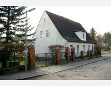 Einfamilienhaus mit weißer Fassade und schwarzem Satteldach an einer Straßenecke, umgeben von einem Zaun mit roten Backsteinpfeilern. Im Vordergrund befinden sich Straßenschilder, die die Kreuzung von Glienicker Straße und Stölpchenweg markieren.