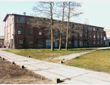 Backstein-Gebäude im Industriestil mit mehreren großen Fenstern und einem Satteldach, umgeben von einer offenen Grünfläche. Im Vordergrund ein gepflasterter Weg und Schienen, die auf das Gebäude zulaufen, sowie mehrere kahle Bäume.