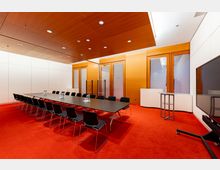 Moderner Konferenzraum mit langem rechteckigem Tisch und schwarzem Stuhlarrangement auf rotem Teppichboden. Die Wände sind in Weiß und Holz gehalten, mit großen Fenstern und Türen mit Holzrahmen sowie einem Flachbildschirm an der rechten Seite.