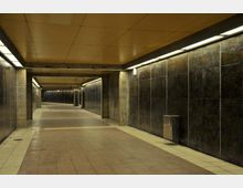 Unterführung mit gelblicher Decke, gefliesten Wänden in dunklem Farbton und einem Boden aus großen, rechteckigen Kacheln. Der Raum wird von länglichen Deckenlampen beleuchtet, und es sind vereinzelt Mülleimer an den Wänden angebracht.