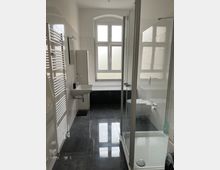 Modernes Badezimmer mit großer, gefliester Dusche, Waschbecken und Toilette. Das Zimmer hat eine große Fensterfront mit satiniertem Glas, einen Heizkörperhandtuchhalter und dunkle, glänzende Bodenfliesen.