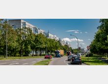 Hauptstraße in einer städtischen Umgebung mit mehrspurigem Verkehr, umgeben von Bäumen und modernen Wohnblocks im Hintergrund. Verkehrsschilder und Straßenlaternen säumen die Straße, während Autos und Lastwagen in beiden Fahrtrichtungen unterwegs sind.