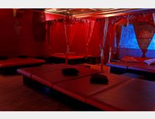 Ein dunkler Raum mit roten Wänden und mehreren roten Liegeflächen, die durch Vorhänge mit roten Stoffen voneinander getrennt sind. Im Hintergrund ist eine metallische Wanddekoration mit Schriftzug und blauer Beleuchtung sichtbar.