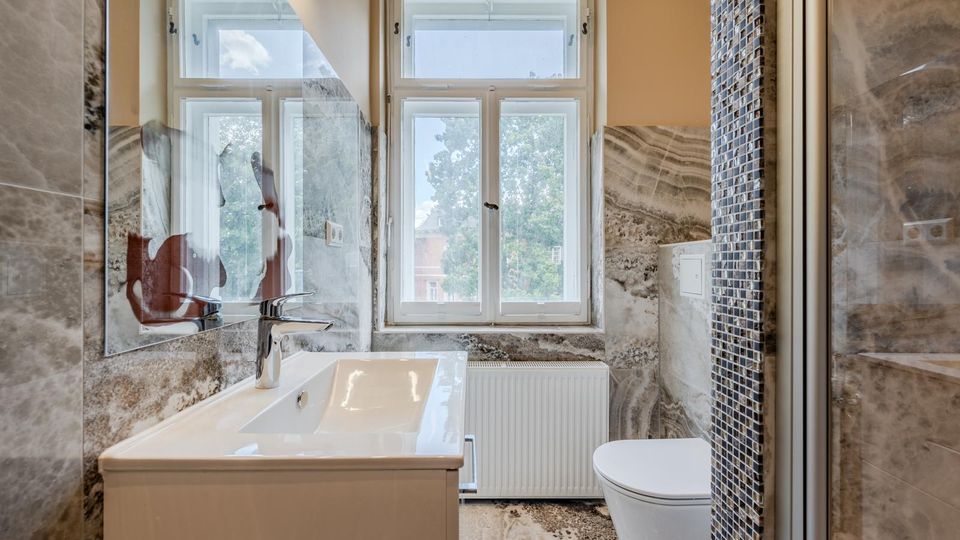 Modernes Badezimmer mit großformatigen marmorierten Wand- und Bodenfliesen, Waschtisch mit Spiegel sowie Toilette neben einer Duschabtrennung mit schmalem Mosaikstreifen. Zwei hohe Fenster über einem Heizkörper lassen Tageslicht herein und bieten Blick ins Grüne.