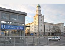 Ansicht des U-Bahnhofs Frankfurter Tor in Berlin mit einem modernen gläsernen Eingangsgebäude im Vordergrund. Im Hintergrund ist ein monumentales Gebäude im neoklassizistischen Stil mit einem markanten grünen Kupferturm zu sehen.