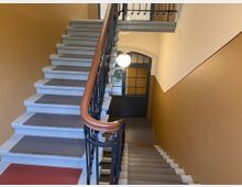 Treppenhaus in einem Altbau mit braun gestrichenen Wänden, weißen Stufen mit grauen und roten Trittschutzflächen und einem kunstvoll verzierten schwarzen Metallgeländer. Im Hintergrund befindet sich eine Doppeltür mit Glaselementen und einem runden Deckenleuchter.