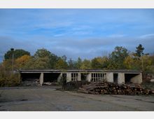 Ein verlassenes, niedriges Gebäude mit offenen Toren und Fenstern, umgeben von Bäumen in herbstlicher Färbung. Im Vordergrund liegt ein Stapel Baumstämme, und die Umgebung wirkt überwachsen und ungenutzt.