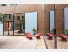 Modernes Gebäude mit einer Glasfassade und Spiegelung angrenzender Architektur. Im Innenhof stehen rote Designerstühle und ein kleiner Tisch auf hellem Bodenbelag, umgeben von Pflanzen in Töpfen.