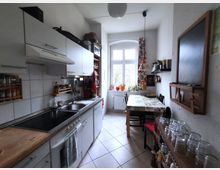 Narrow galley kitchen with white cabinets and tiled backsplash along one wall, a long countertop with sink and stove, and a small dining table with chairs on the opposite side. A tall arched window at the end lets in daylight, with potted plants on the sill, patterned curtains, wall shelves, and a chalkboard.