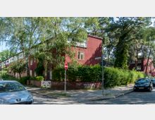 Eine Straßenecke in einem Wohngebiet mit mehrstöckigen roten Backsteinhäusern, umgeben von Bäumen und Hecken. Der gepflasterte Gehweg ist teils von parkenden Autos gesäumt, und Straßenschilder markieren die Kreuzung von "Am Fuchspfad" und "Hodichsweg".